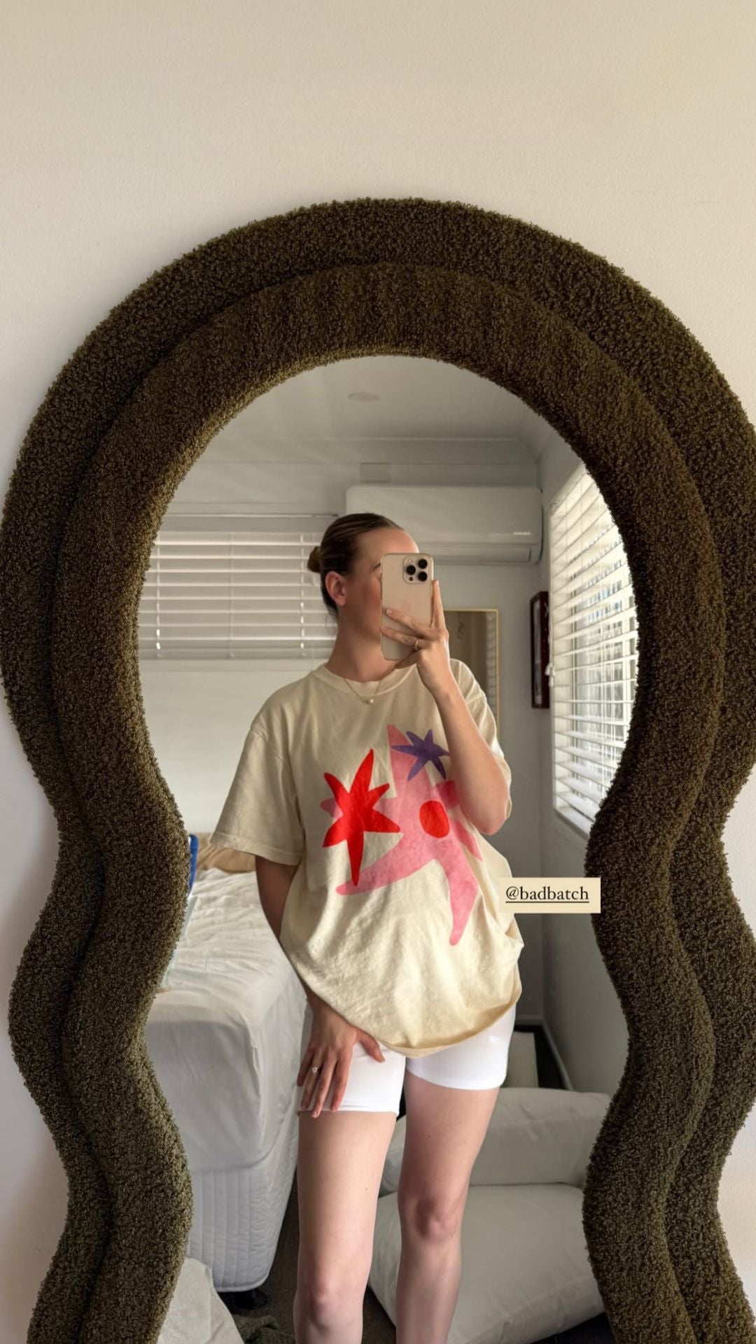 Person taking a mirror selfie wearing a beige t-shirt with a colorful design and white shorts, standing in a room with a textured mirror.