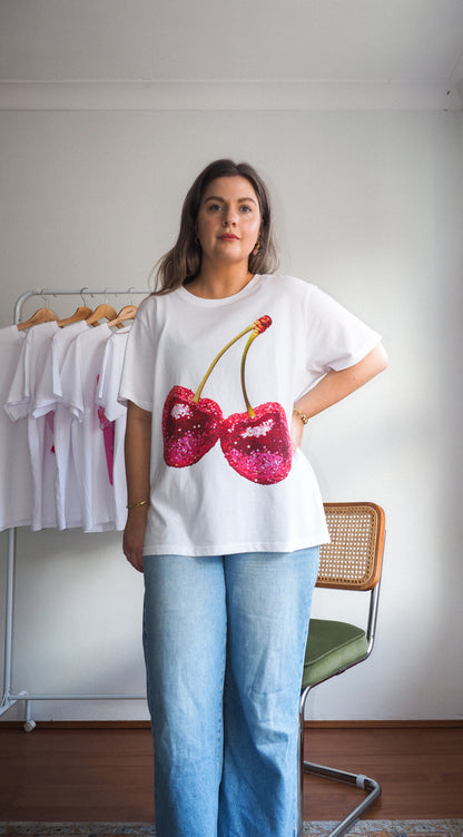Woman modeling white tee with large pink sequin cherries, styled with light blue jeans, standing in front of a t-shirt rack in a minimal room